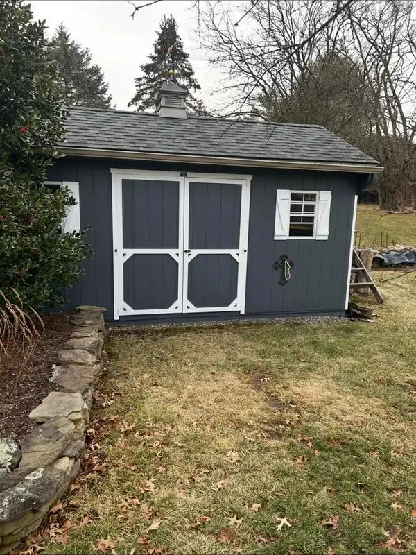 Newly built dark gray storage shed with white trim, barn-style double doors and cupola weathervane, beside a stone garden wall.