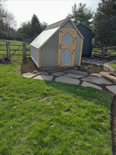 Wooden garden shed with a stone paver path leading to it, set in a mulch bed, rail fence behind.