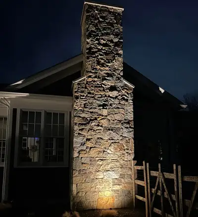 Night: stone chimney uplit by landscape lighting, wood fence and gate beside it.