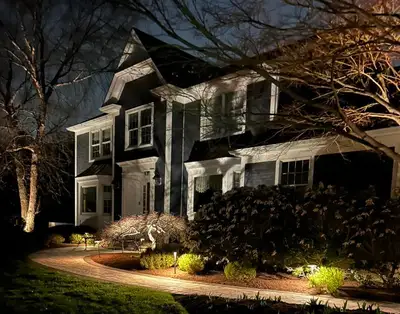 Night: two-story house facade lit by landscape lighting, curved paver walkway, planting beds, bench near the entry.