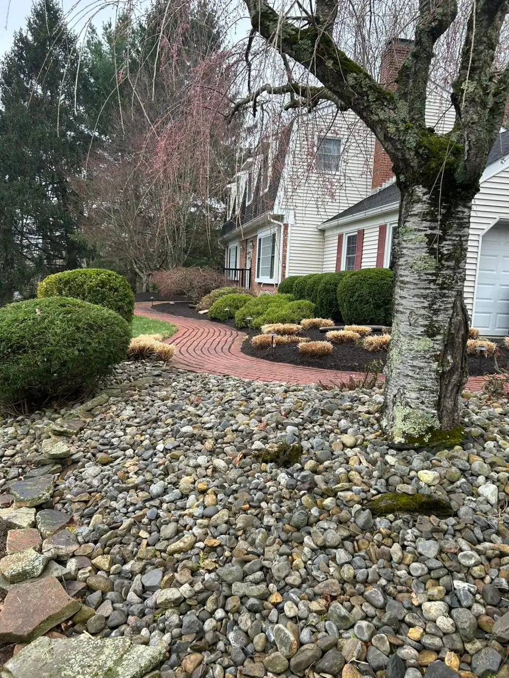 Curved brick walkway with mulched foundation beds, river-rock border and trimmed shrubs, weeping tree in foreground.