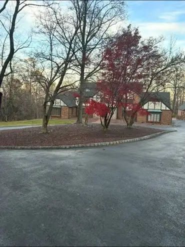 Late fall cleanup with fresh mulch ring by driveway, bare trees, red Japanese maple, brick house.