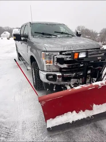 A silver RFW pickup truck with a red snowplow blade sits on a snow-covered lot under an overcast sky.