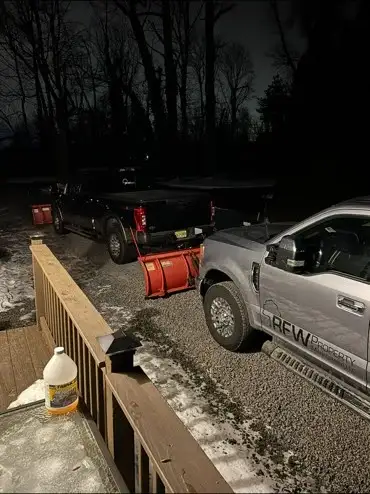 Two RFW pickup trucks with snowplows parked at a house at night, light snow on ground.
